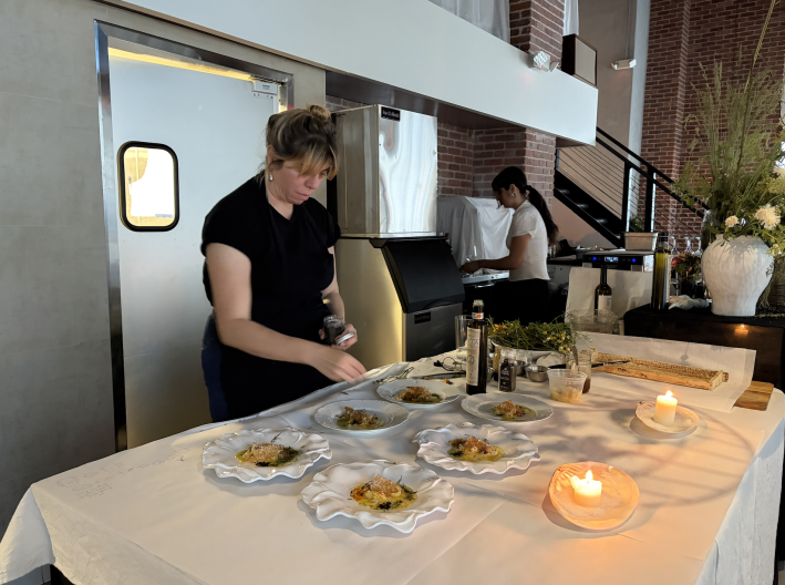 A light skinned woman with her blonde hair up, wearing a black T shirt and pants, is bending over a table, sprinkling something over six plates full of food.