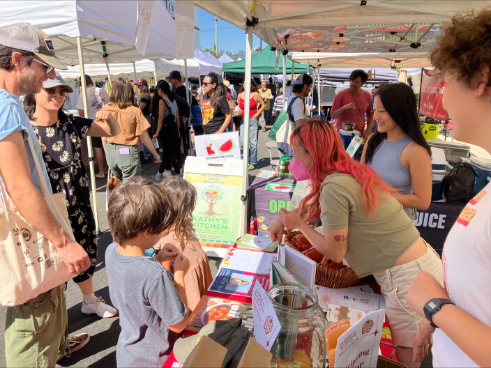 A woman with a mask and orange hair leaning showing children a book under at a tent in a farmers' market.