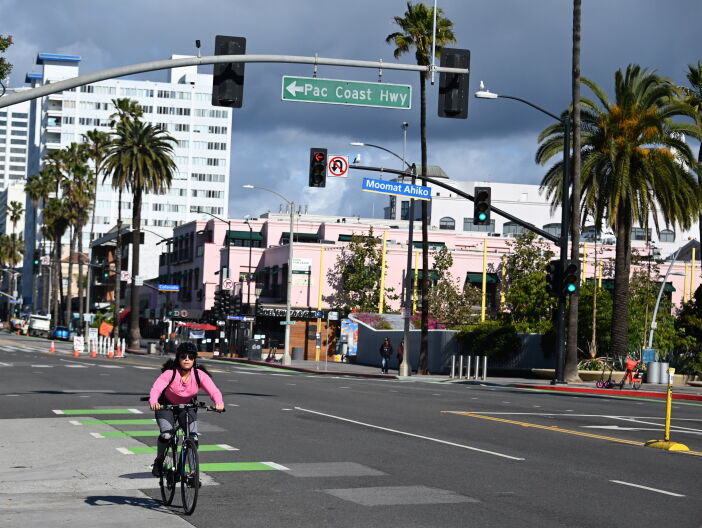 A woman in a pink shirt wearing a backpack rides a bike down a road in Santa Monica. Palm trees and buildings are in the background. A street sign behind her marks the Pacific Coast Highway.