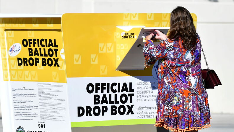 A person puts her ballot through the slot of an official drop box. The ballot box is orange and white with large black letters, reading "official ballot drop box."