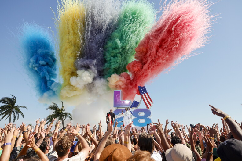 A colorful firework explodes in the sky in blue, yellow, purple, green and red colors. A sign that reads "LA28" is on a stand before a crowd of people.