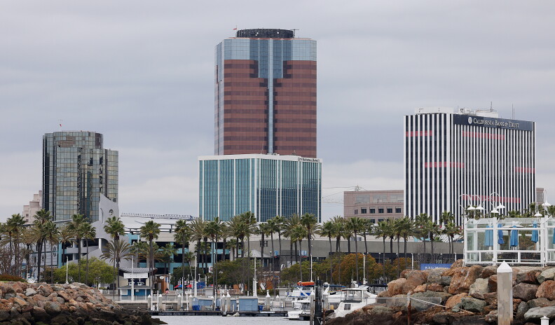 Long beach skyscrapers in front of a cloudy sky. A row of palm trees line the front at the bottome of the buildings.