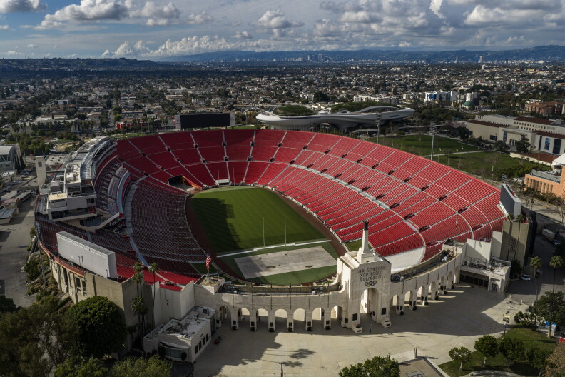 An aerial view of audience stands and a grassy field. Buildings are in the distance behind the arena.