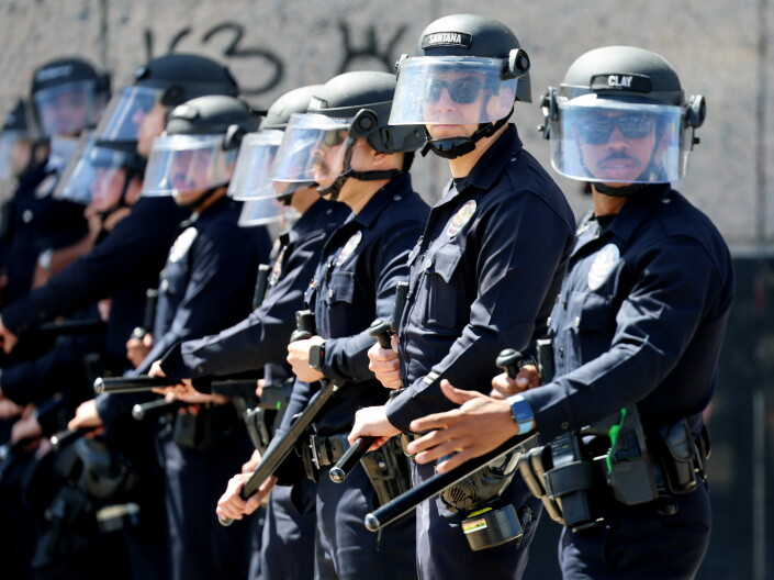 A man with medium skin ton and a bald head appears to be filming an ICE officer in a camouflage uniform.