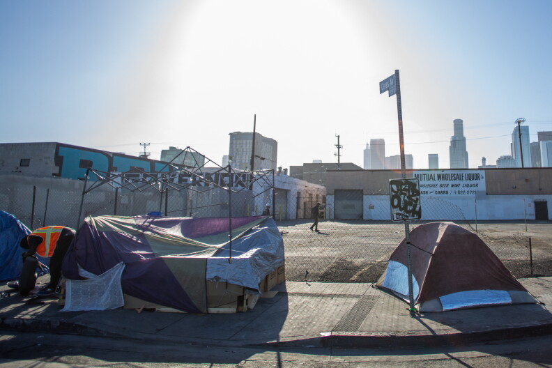 Tents are erected on sidewalk next to a chainlink fence that surround a warehouse. A downtown skyline is in the distance.