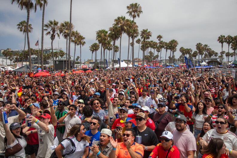 A large crowd of people are cheering and smiling towards a screen seen behind the picture frame. Palm trees are in the distance with multi-colored tents.