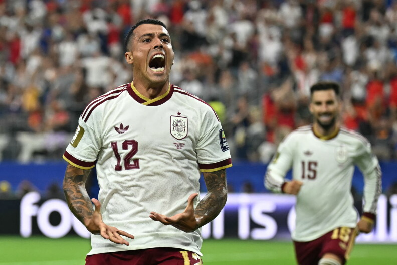 A soccer player wearing a white and burgundy soccer jersey celbrates on a soccer field. A team player runs towards him from behind.