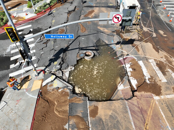 An aerial view of a street intersection: A portion of the street has caved in, exposing a sink hole swirling with muddy waters. 