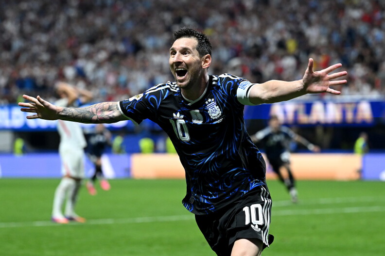 A soccer player wearing a blue jersey, runs across a soccer field celebrating with his arms stretched out.