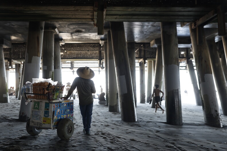 Man wearing a straw hat leans on an ice cream cart in the shade of a pier on a beach.