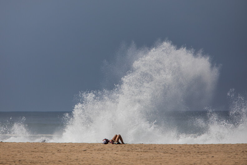 A woman sunbathes on the beach as a big wave hits the breakwater in Venice Beach, sending white spray into the air.