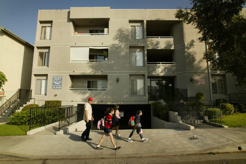 A man walks with three school-aged girls past a multi-story tan apartment building. The numbers "1051" are displayed in white on the front over a doorway. Patches of green grass and small shrubs are seen on either side of the building.
