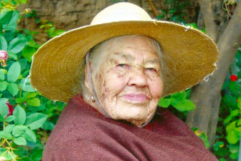 A woman wearing a wide-brimmed straw hat and a maroon shawl, smiles at the camera in a garden.
