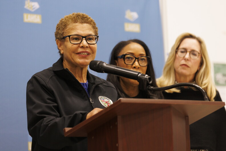 A woman with medium skin tone with short curly light brown hair wearing black-rimmed glasses and a black jacket with the seal of Los Angeles stands behind a podium speaking into a microphone.