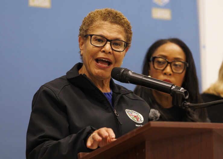 A woman with medium skin tone with short curly light brown hair wearing black-rimmed glasses and a black jacket with the seal of Los Angeles stands behind a podium speaking into a microphone. 