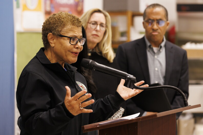 A woman with medium skin tone with short curly light brown hair wearing black-rimmed glasses and a black jacket with the seal of Los Angeles stands behind a podium speaking into a microphone.