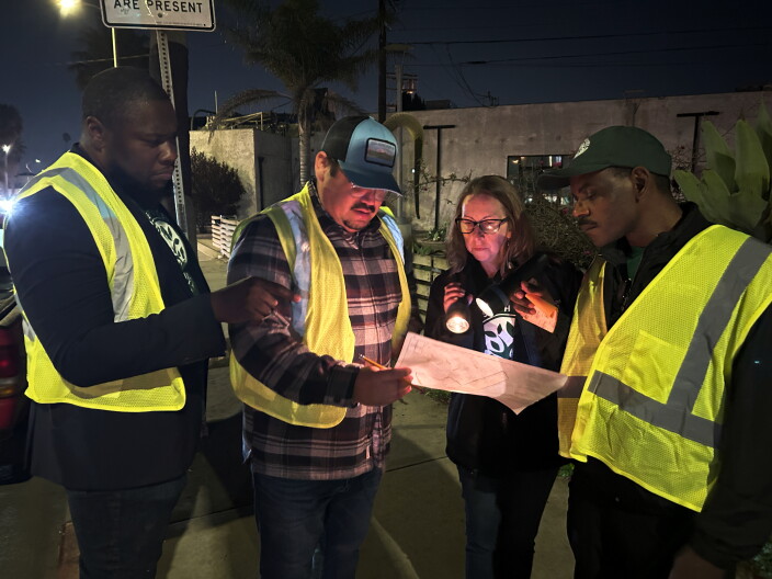 4 people gather around a map at night. 3 are wearing yellow reflective vests, and the other is shining a flashlight.