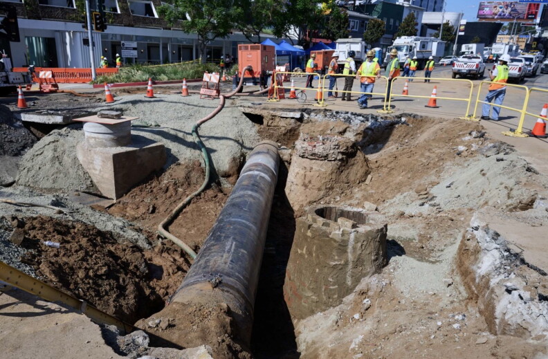 A section of a city street has been dug up, creating a deep trench with an exposed pipe running down the middle. 