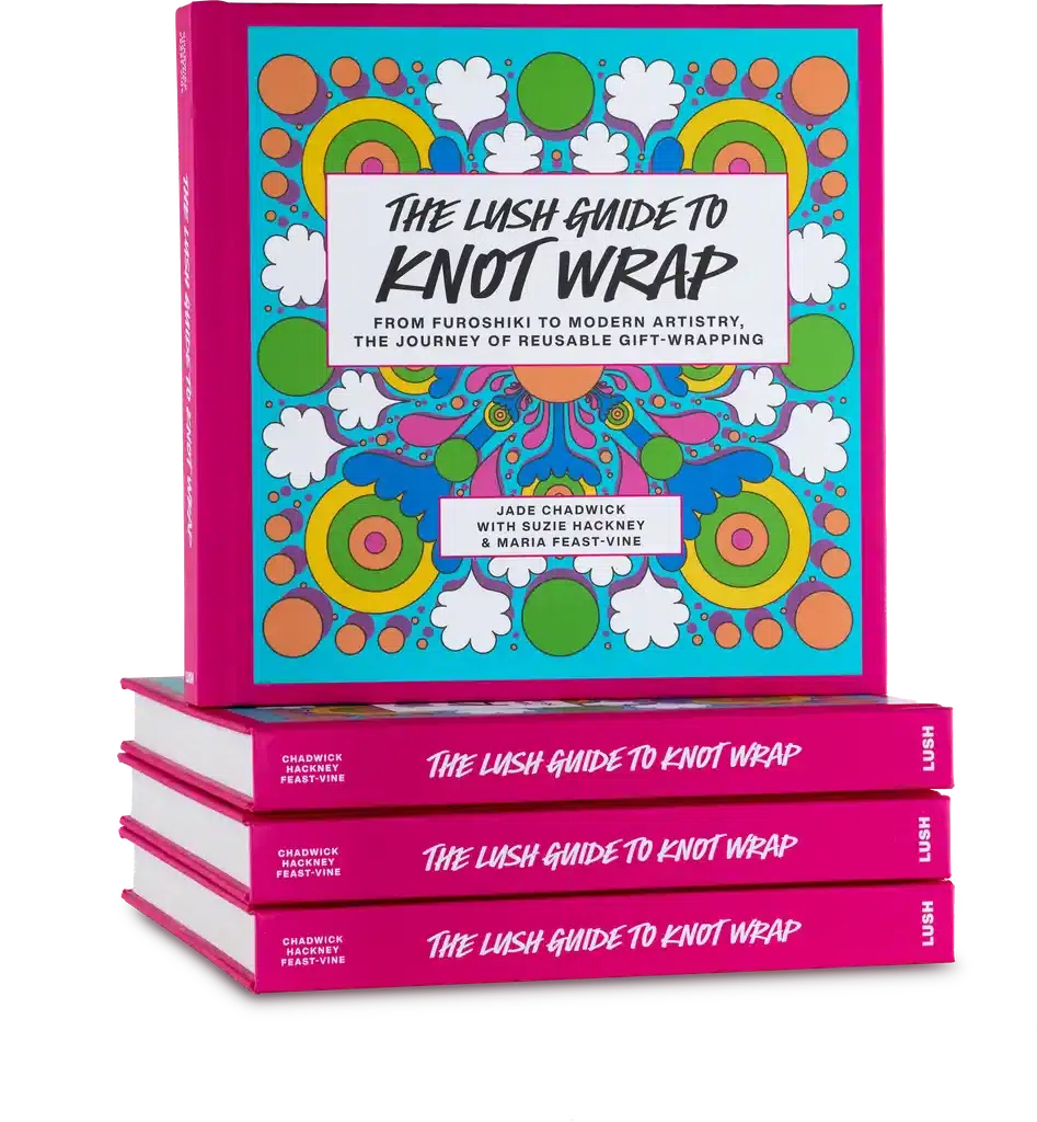 A display of "The LUSH Guide to Knot Wraps". The main book on the top is bright pink with a psychedelic green, blue and orange Knot Wrap on the front. It is displayed on top of three other copies of the book.