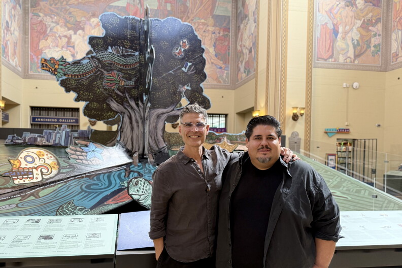 Two men pose in front of a giant pop-up-book art installation featuring a tree, a feathered serpent and a sea turtle inside the LA Central Library rotunda.