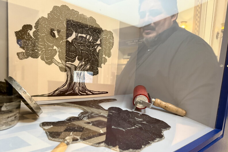 A linocut print of an oak tree sits in a display case alongside the carved block, ink roller and carving tools used to make it.