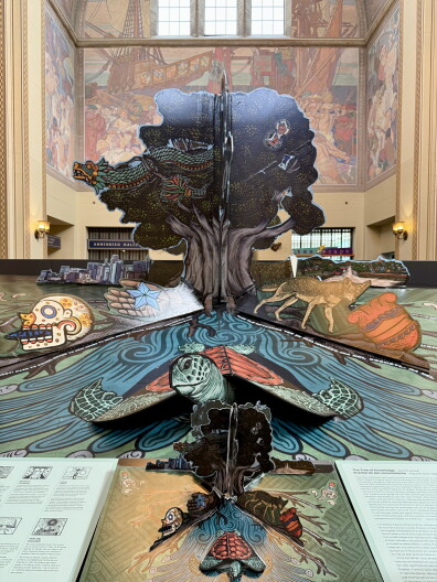 A giant pop-up spread featuring a tree, feathered serpent, coyote and sea turtle towers over a regular-sized copy of the same pop-up book at the L.A. Central Library.