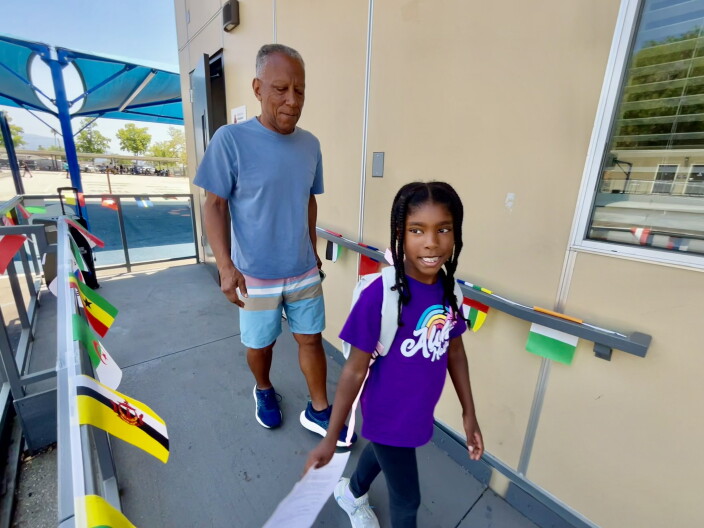 An eight-year-old Black girl is trailed by her grandfather going past school trailers.