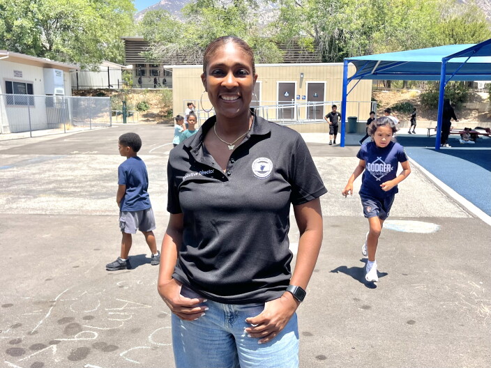 A Black woman in a black polo shirt and jeans poses in front of an outdoor school play area as several children play in the background.