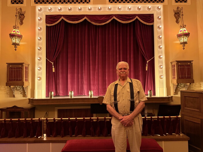 A man wearing suspenders stands in front of a stage with a red carpet.