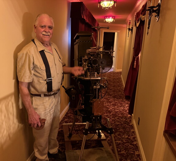 An older man with a gray mustache wearing a tan shirt, white pants and suspenders is handcranking an antique movie projector