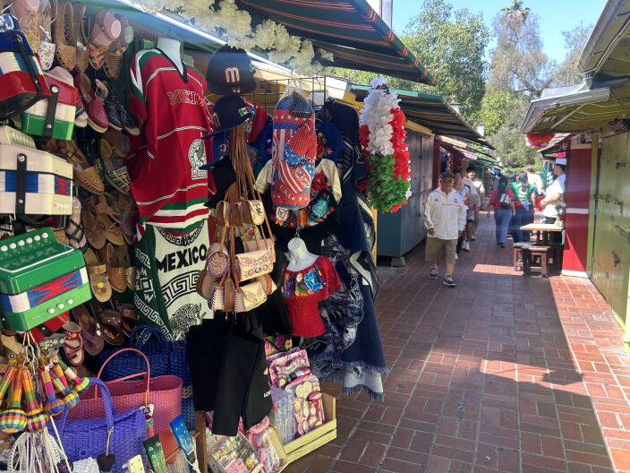 Outside a shop hangs a variety of items, including colorful ponchos, toy accordions, and leather bags. Surrounding shops are closed. A few people walk through the cobblestone street.
