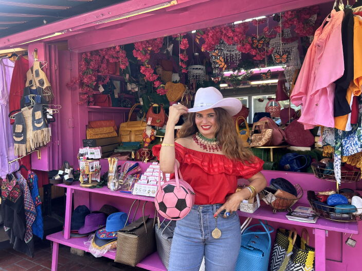 A woman - wearing a white tejana, red blouse and a red, white and green beaded necklace - stands in front of her hot pink shop. Her kiosk is adorned with hats, bags and clothing items. She's holding a small pink handbag designed to look like a soccer ball.