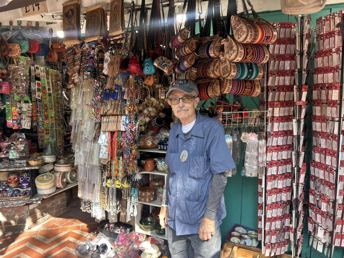 An older man, wearing glasses, a baseball cap and a blue button-up, stands in front of his store. He's surrounded by his many products which include leather bags, jewelry, and home decor.