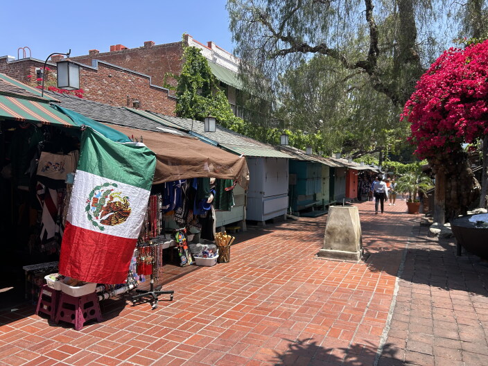 Wooden kiosks line a cobblestone street. A Mexican flag hangs off the shade of the kiosk at the front. A person at a distance walks away from the shops.