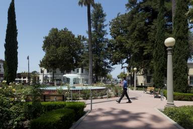 A man wearing black pants and a blue long sleeve walks across a plaza with a large water fountain at its center. 
