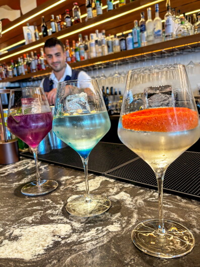 Three colored gin tonics on a bar top with a bartender standing behind a wall of bottles.