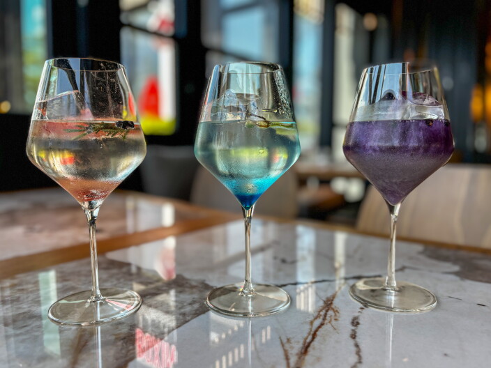 Three gin tonics in stemmed glasses on a marble table, garnished with rosemary and shifting from clear to blue to deep purple.