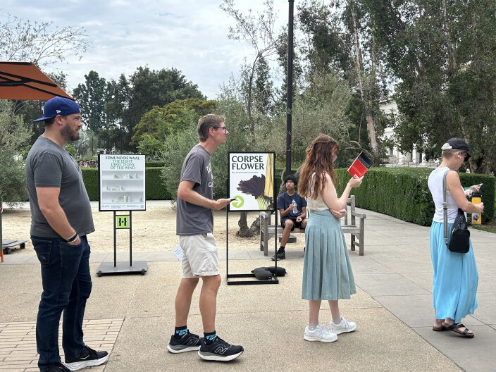 Four people, two men and two women, are lined up single file in front of a sign that reads "CORPSE FLOWER."
