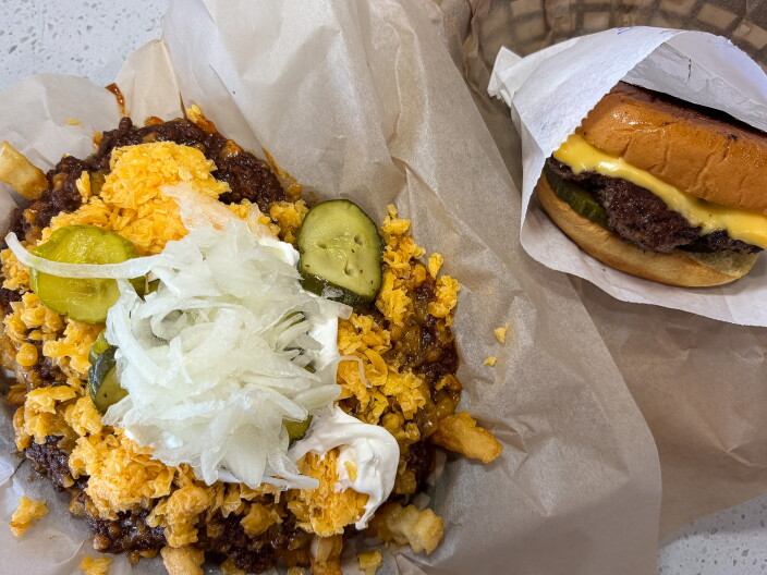 A chili cheese burger wrapped in white paper sits next to a basket of chili cheese fries topped with shredded cheddar, sour cream, raw onion, and pickles on white parchment paper.
