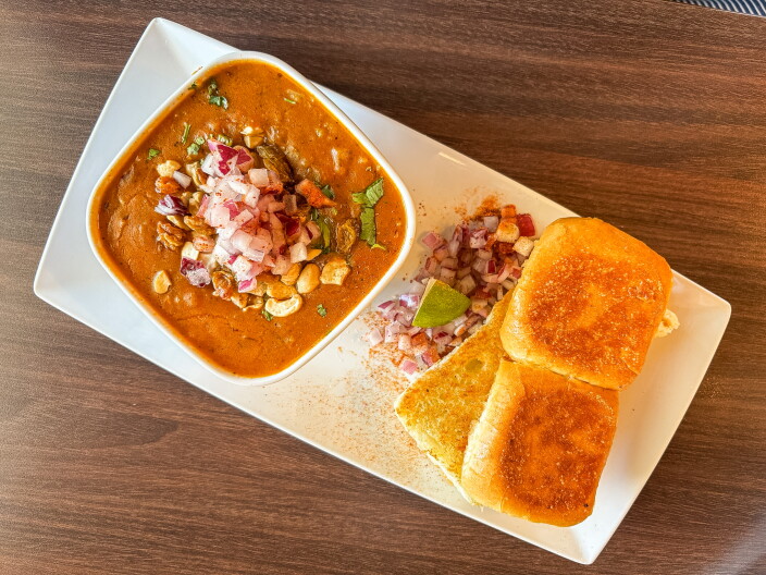 An overhead shot of the Honest Special Bhaji Pav β a rich orange-red spiced curry topped with cashews, raisins, and diced red onion in a white bowl, served alongside two golden toasted pav rolls on a white rectangular plate.