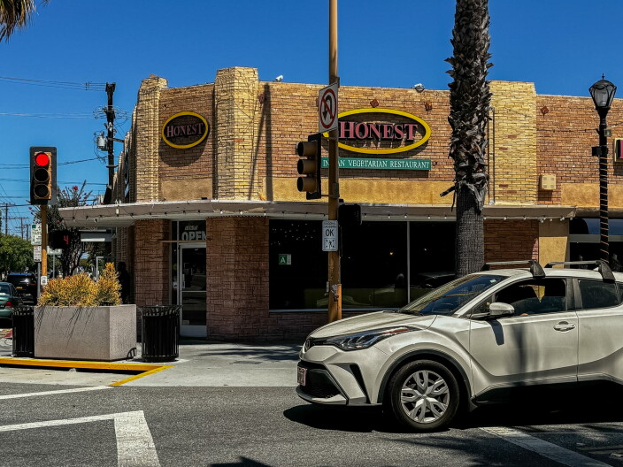 The brick exterior of Honest Indian Vegetarian Restaurant on a corner lot on Pioneer Boulevard, with two yellow-rimmed oval signs reading "Honest" above a green banner reading "Indian Vegetarian Restaurant." There's also a traffic light in the foreground and palm trees visible against a blue sky.
