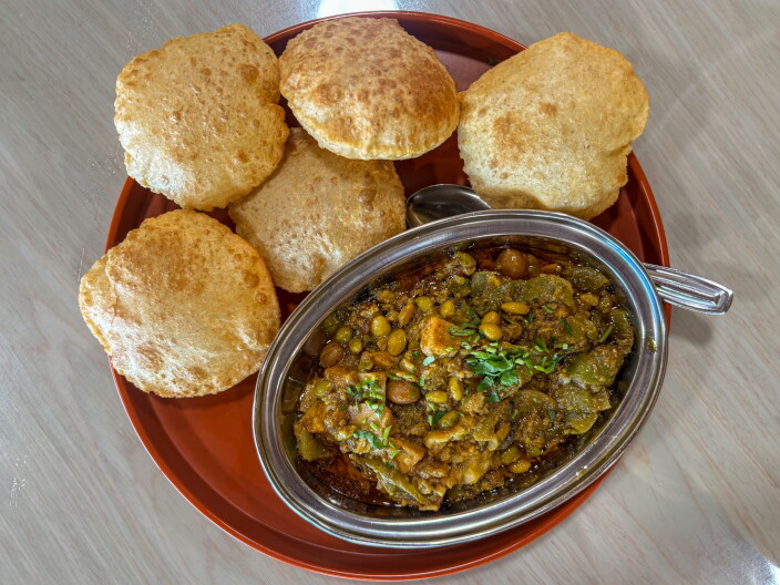 An overhead shot of the Undhiyu Puri at Jay Bharat β a dark, complex Gujarati vegetable curry with visible legumes and a fresh herb garnish in a silver kadai, surrounded by five golden puffed puris on a red tray