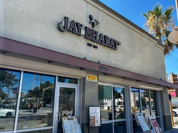 The exterior of Jay Bharat restaurant at 18701 Pioneer Boulevard in Artesia, with bold black three-dimensional lettering and "Est. 1985" below the name, an Open neon sign glowing yellow above the entrance, and menus displayed outside on a sunny day.