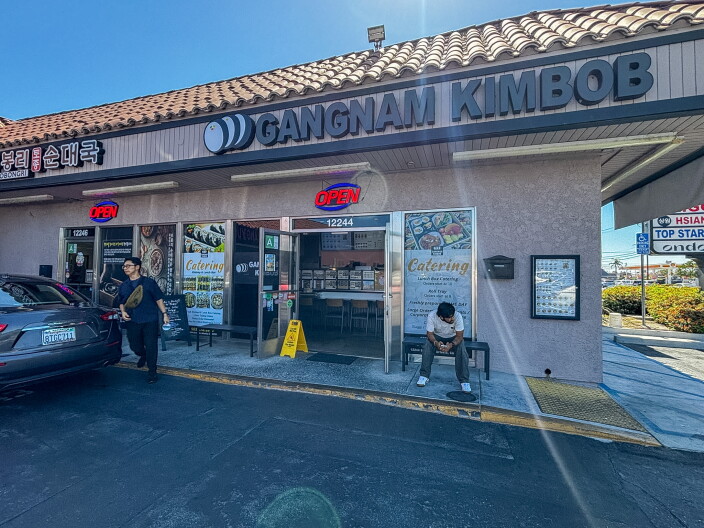 The exterior of Gangnam Kimbob with large block lettering on the facade, two Open signs in the windows, catering signage on the door, and a person sitting outside on a sunny day.