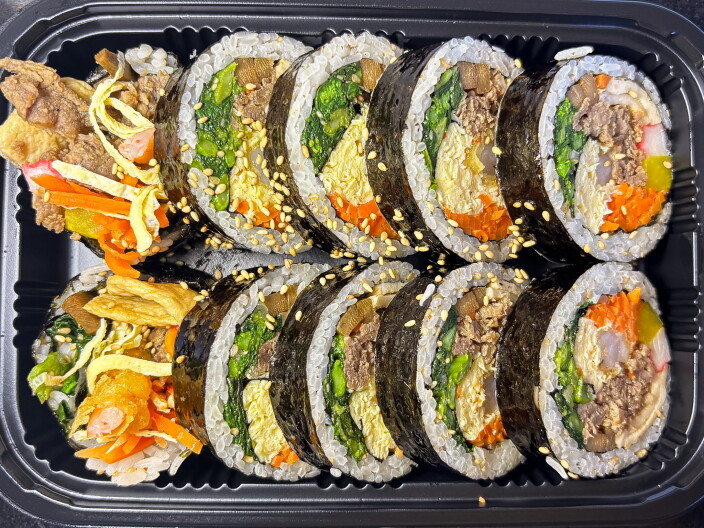 An overhead shot of ten pieces of kimbob arranged in two rows in a black plastic container, showing colorful cross-sections of bulgogi beef, spinach, egg, carrot, and pickled vegetables wrapped in nori and sesame-oil rice, topped with sesame seeds.