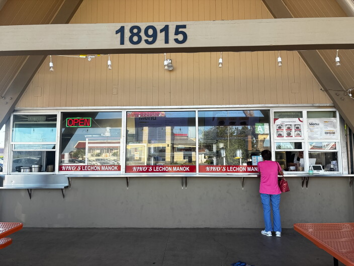 A customer in a pink shirt orders at the window counter of Kiko's Lechon Manok, a Filipino rotisserie chicken spot housed in a large A-frame building with red patio tables visible in the foreground.