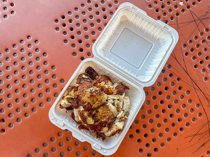 An overhead shot of a styrofoam container filled with pieces of rotisserie chicken with dark brown crispy skin, sitting open on an orange perforated metal patio table at Kiko's Lechon Manok.