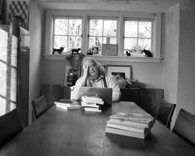 A male presenting person sits at a wood table. There are three stacks of books in front of him.