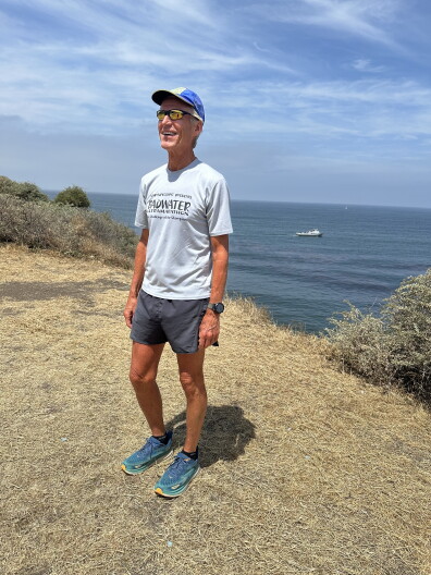 A man in a hat and sunglasses standing in runner shorts and a t-shirt in front of the ocean.