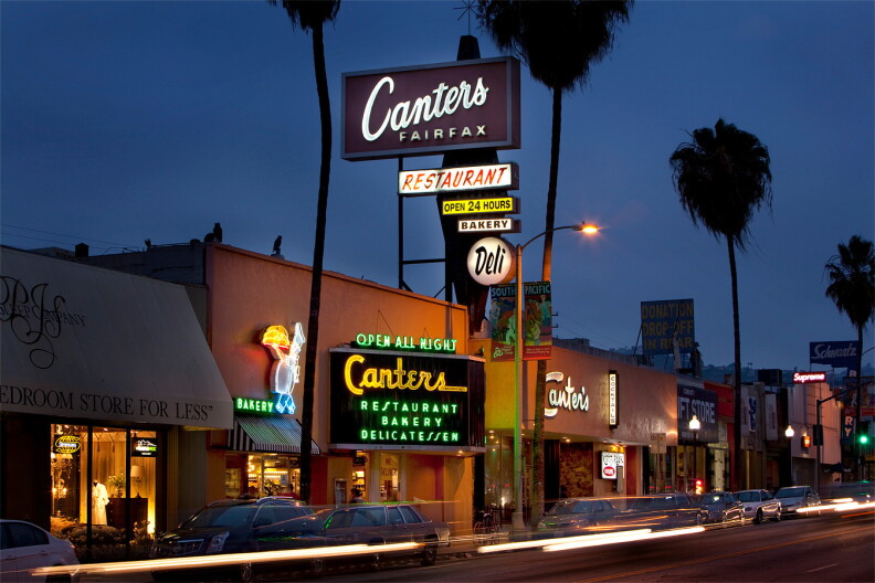 Canter's Deli's neon sign glows above Fairfax Avenue at dusk, reading "Restaurant," "Open 24 Hours," and "Bakery," with palm trees and passing car lights in the foreground.